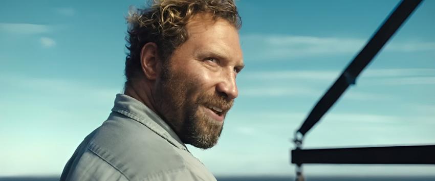 Dangerous Animals (1970) Movie scene: Bruce Tucker (Jai Courtney), rugged with curly blonde hair and a beard, glances back with his winch visible against a bright blue sky backdrop