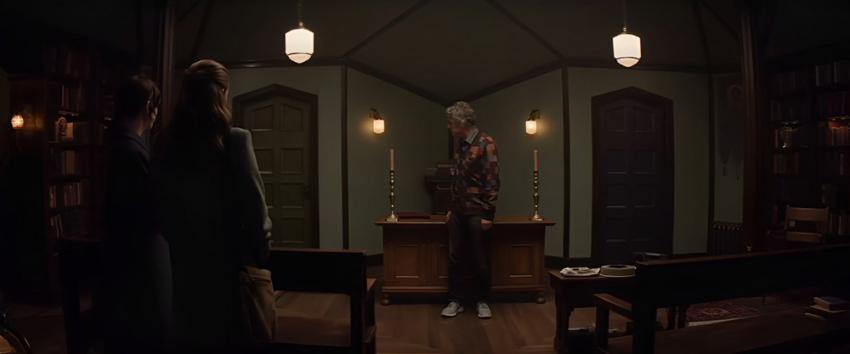 Heretic (1970) Movie scene: Mr. Reed (Hugh Grant) stands at a wooden desk, flanked by two missionaries, Sister Barnes (Sophie Thatcher) and Sister Paxton (Chloe East)