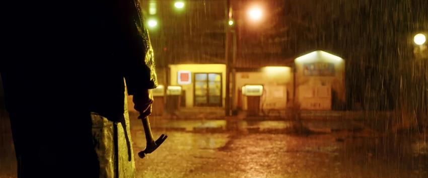 Open 24 Hours (1970) Movie scene: A shadowy figure holding a hammer stands in the rain at night, facing a brightly lit secluded gas station