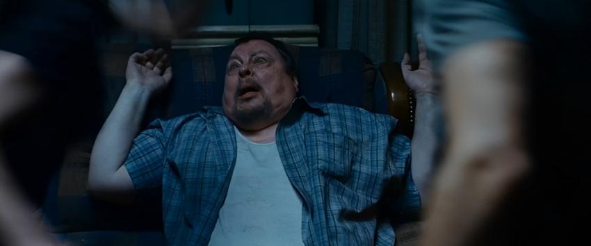 Pathology (1970) Movie scene: A man in a blue plaid shirt and white undershirt appears panicked and raises his arms while sitting on a couch.