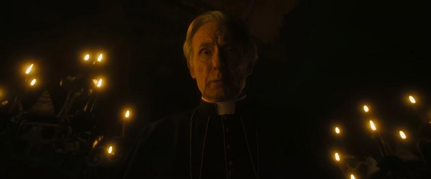 The First Omen (1970) Movie scene: Cardinal Lawrence (Bill Nighy) standing amongst candles, gazing downward, enveloped in worry and contemplation.