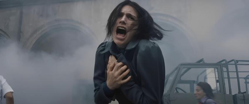 The First Omen (1970) Movie scene: Margaret (Nell Tiger Free), in a dark coat, screams in terror with hands on her chest, as fog swirls around her in a chaotic street scene with stone arches and a military vehicle, evoking the unrest of 1970s Rome.