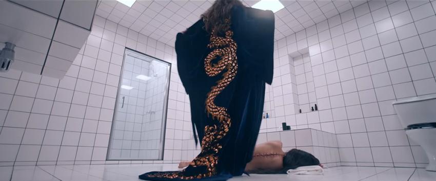 The Substance (1970) Movie scene: A person, seen from behind, is standing and wearing a dark robe adorned with a golden dragon design in a white-tiled bathroom