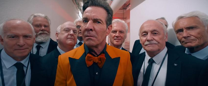 The Substance (1970) Movie scene: Harvey (Dennis Quaid) stands at the center of a group, wearing a vibrant orange suit jacket and sporting a confused expression