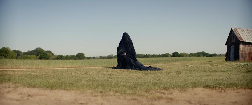 The Woman in the Yard (1970) Movie scene: Front-facing and seated in the yard is the black-clad woman in the yard with a veil covering her face.